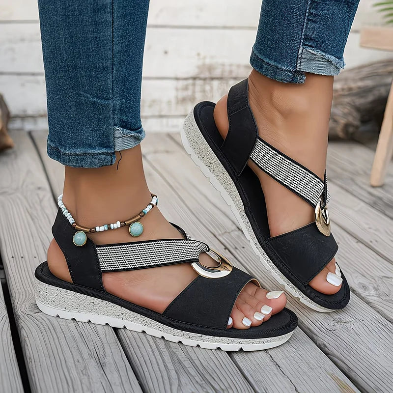 Artemis sandals in black, worn while walking on a paved path.