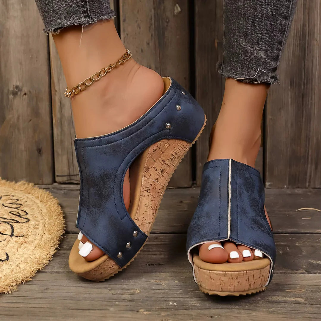 A model wearing beige Solange studded wedge sandals, styled with a flowing summer dress in a garden setting.